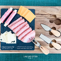 Acacia wood charcuterie board with slate serving surface and three wood-handled serving utensils. Engraved in the style of a dictionary entry stating "Charcuterie a french term for assembling cheese and other fancy shit." Engraved and sold by Like No Otter Design Co.