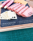 Acacia wood charcuterie board with slate serving surface and three wood-handled serving utensils. Engraved in the style of a dictionary entry stating "Charcuterie a french term for assembling cheese and other fancy shit." Engraved and sold by Like No Otter Design Co.