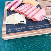 Acacia wood charcuterie board with slate serving surface and three wood-handled serving utensils. Engraved in the style of a dictionary entry stating "Charcuterie a french term for assembling cheese and other fancy shit." Engraved and sold by Like No Otter Design Co.