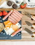Acacia wood charcuterie board with slate serving surface and three wood-handled serving utensils. Engraved in the style of a dictionary entry stating "Charcuterie a french term for assembling cheese and other fancy shit." Engraved and sold by Like No Otter Design Co.