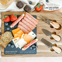 Acacia wood charcuterie board with slate serving surface and three wood-handled serving utensils. Engraved in the style of a dictionary entry stating "Charcuterie a french term for assembling cheese and other fancy shit." Engraved and sold by Like No Otter Design Co.