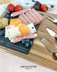 Acacia wood charcuterie board with slate serving surface and three wood-handled serving utensils. Engraved in the style of a dictionary entry stating "Charcuterie a french term for assembling cheese and other fancy shit." Engraved and sold by Like No Otter Design Co.