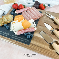 Acacia wood charcuterie board with slate serving surface and three wood-handled serving utensils. Engraved in the style of a dictionary entry stating "Charcuterie a french term for assembling cheese and other fancy shit." Engraved and sold by Like No Otter Design Co.