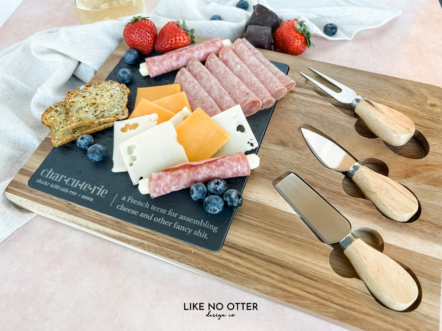 Acacia wood charcuterie board with slate serving surface and three wood-handled serving utensils. Engraved in the style of a dictionary entry stating "Charcuterie a french term for assembling cheese and other fancy shit." Engraved and sold by Like No Otter Design Co.