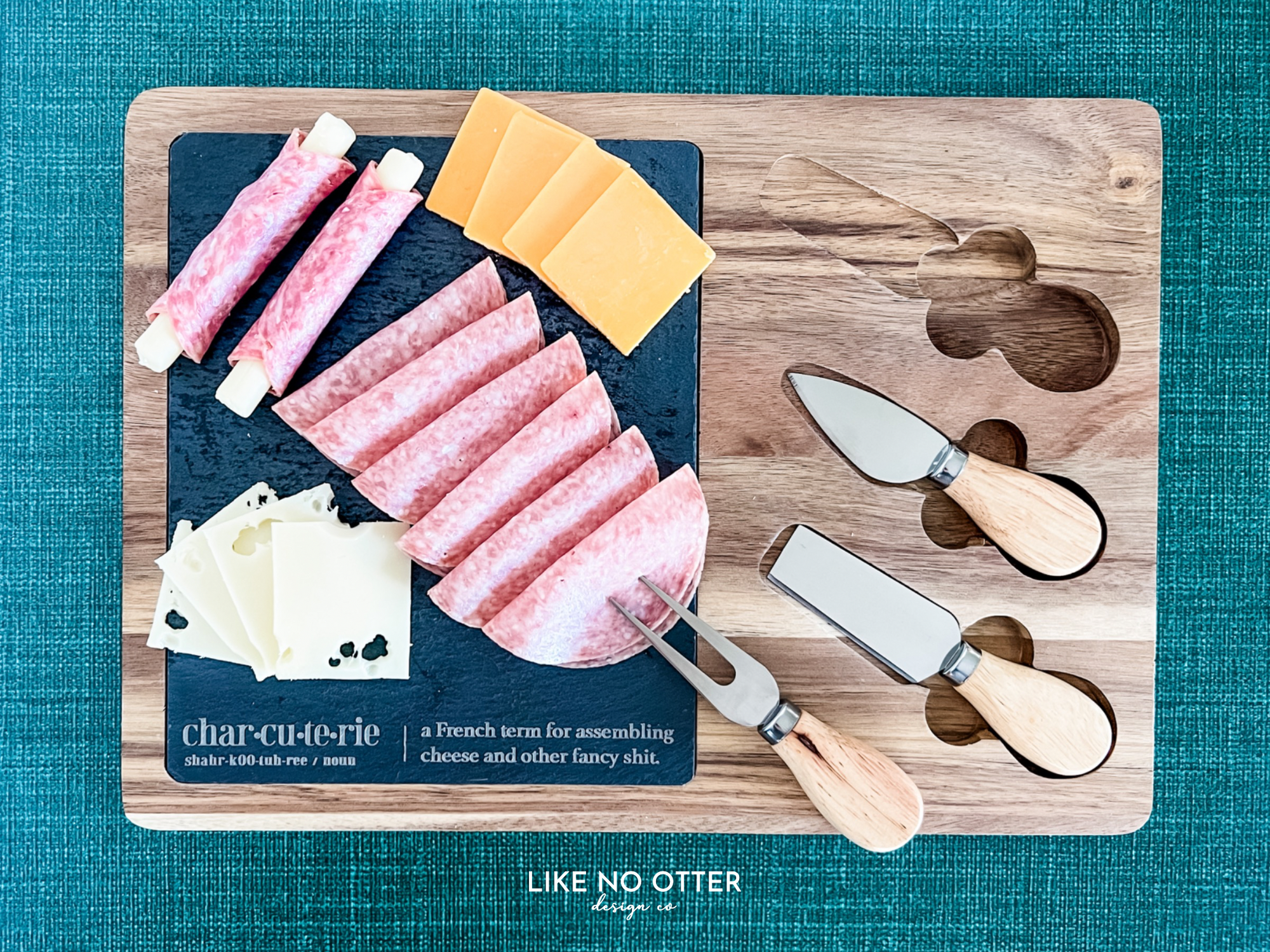 Acacia wood charcuterie board with slate serving surface and three wood-handled serving utensils. Engraved in the style of a dictionary entry stating "Charcuterie a french term for assembling cheese and other fancy shit." Engraved and sold by Like No Otter Design Co.
