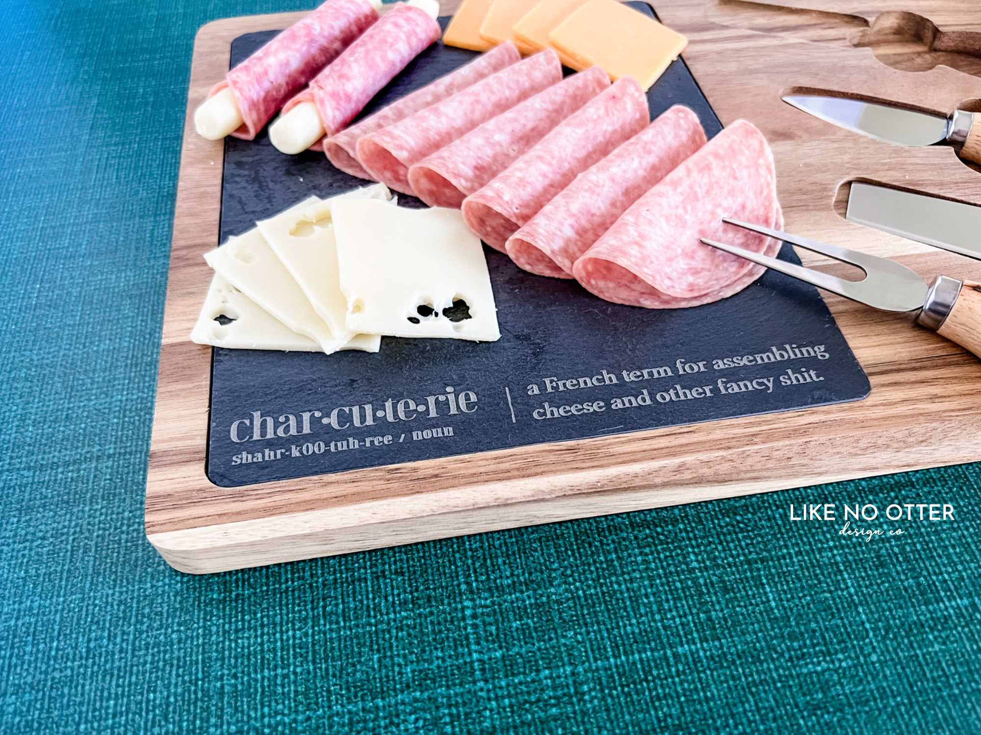 Acacia wood charcuterie board with slate serving surface and three wood-handled serving utensils. Engraved in the style of a dictionary entry stating "Charcuterie a french term for assembling cheese and other fancy shit." Engraved and sold by Like No Otter Design Co.