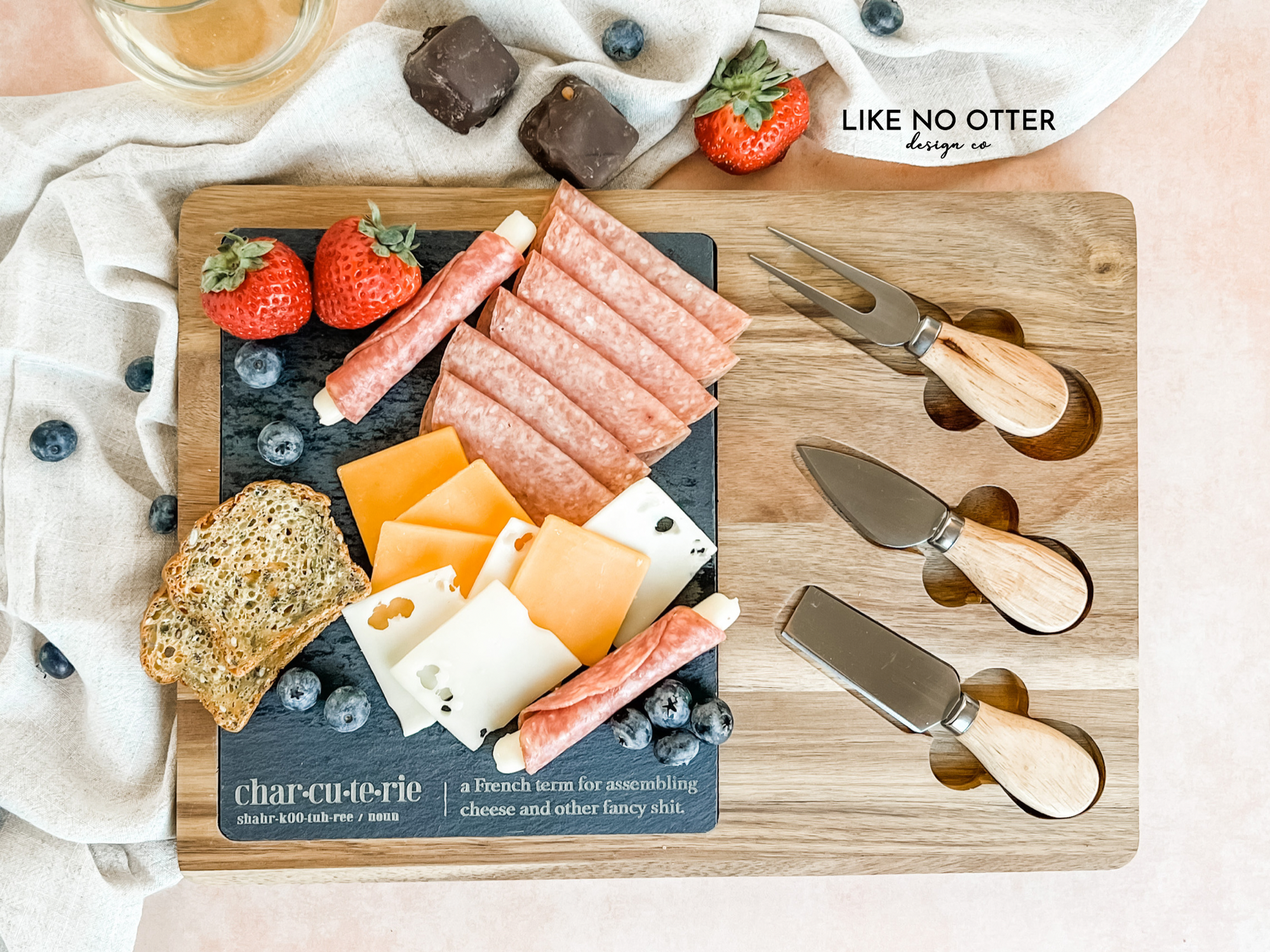 Acacia wood charcuterie board with slate serving surface and three wood-handled serving utensils. Engraved in the style of a dictionary entry stating "Charcuterie a french term for assembling cheese and other fancy shit." Engraved and sold by Like No Otter Design Co.